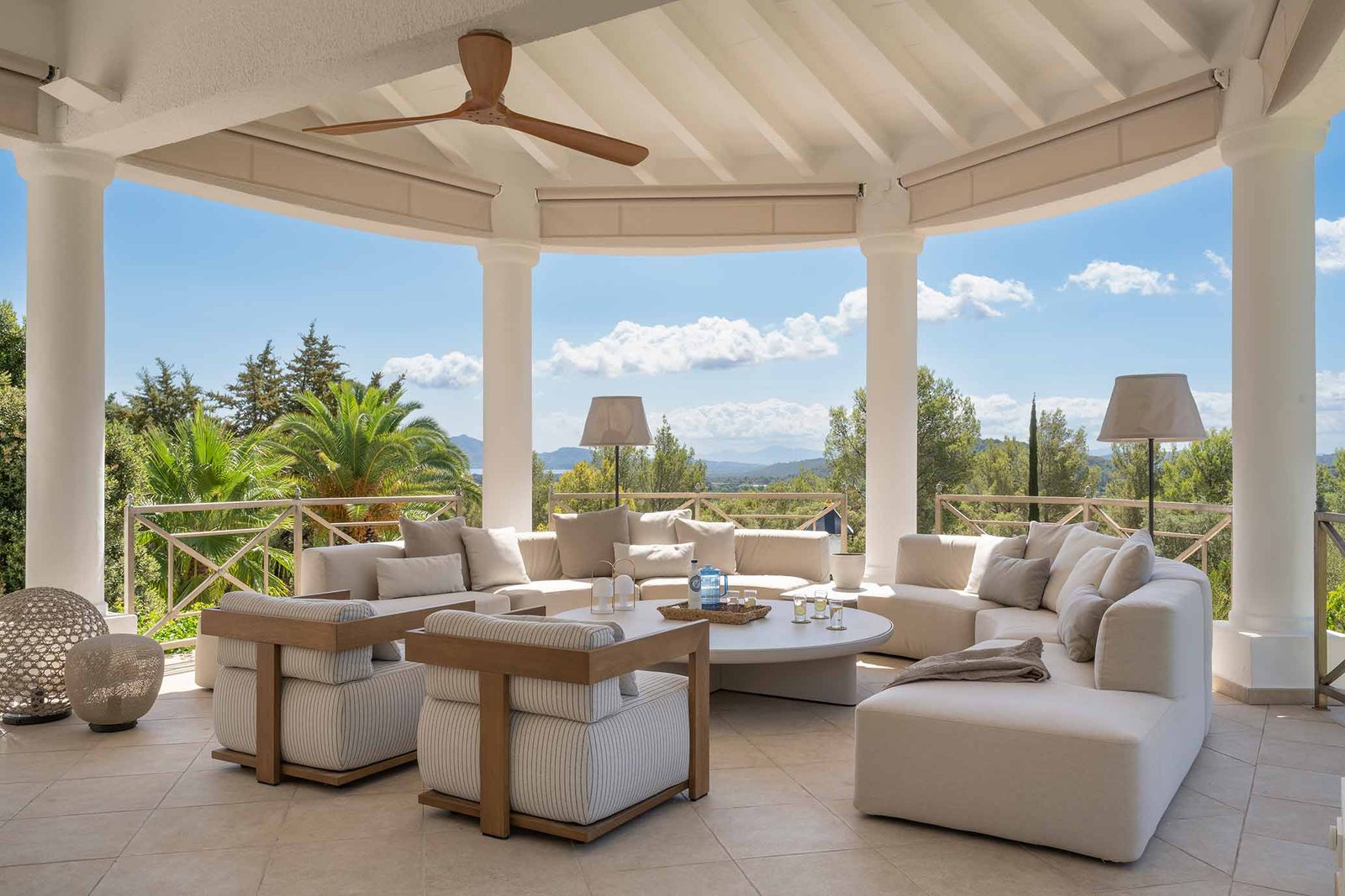 Porche circular de columnas blancas con sofás beige, butacas y vistas a palmeras, pinos y montañas