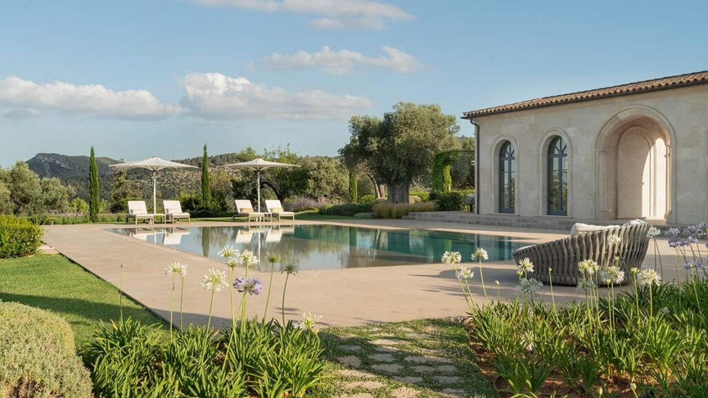 Piscina con tumbonas y sombrillas junto al pabellón de piedra, agapantos en flor y montañas al fondo