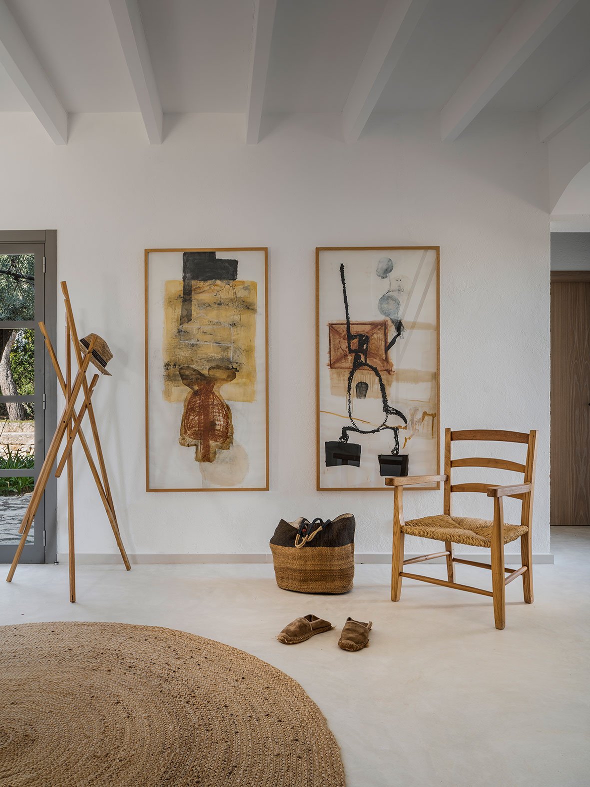 Vestíbulo blanco con dos cuadros abstractos, silla de enea, alfombra de yute y pavimento continuo claro