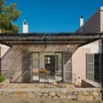 Fachada con pérgola metálica de cañizo, contraventanas grises, terraza de piedra, muro de mampostería y sillas metálicas