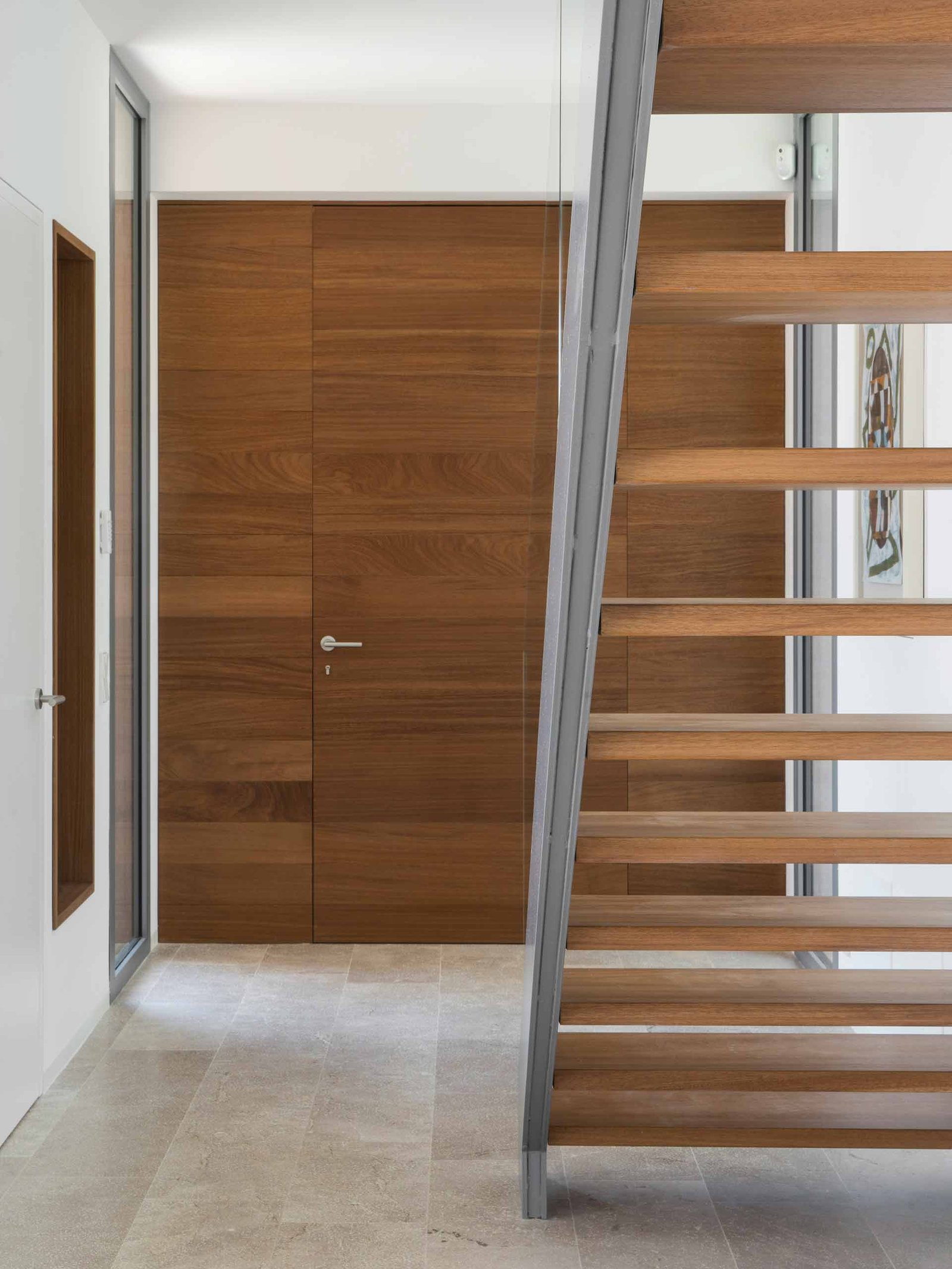Vestíbulo con puerta enrasada de madera, suelo de piedra clara y escalera de peldaños de madera