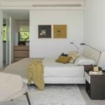 Dormitorio con cabecero tapizado, dos cuadros abstractos, cómoda de madera al fondo y suelo de piedra