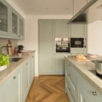 Cocina en verde salvia con encimera de piedra clara, horno empotrado, isla y suelo de parqué en espiga