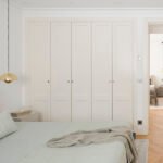 Dormitorio con colcha verde suave y manta gris, puerta abierta al pasillo y otra habitación al fondo