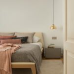Dormitorio con cama de madera y cabecero beige, mantas rosas y grises, mesita gris y lámpara de latón