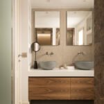 Baño con doble lavabo gris sobre mueble de madera, dos espejos, pared de piedra beige y mármol marrón