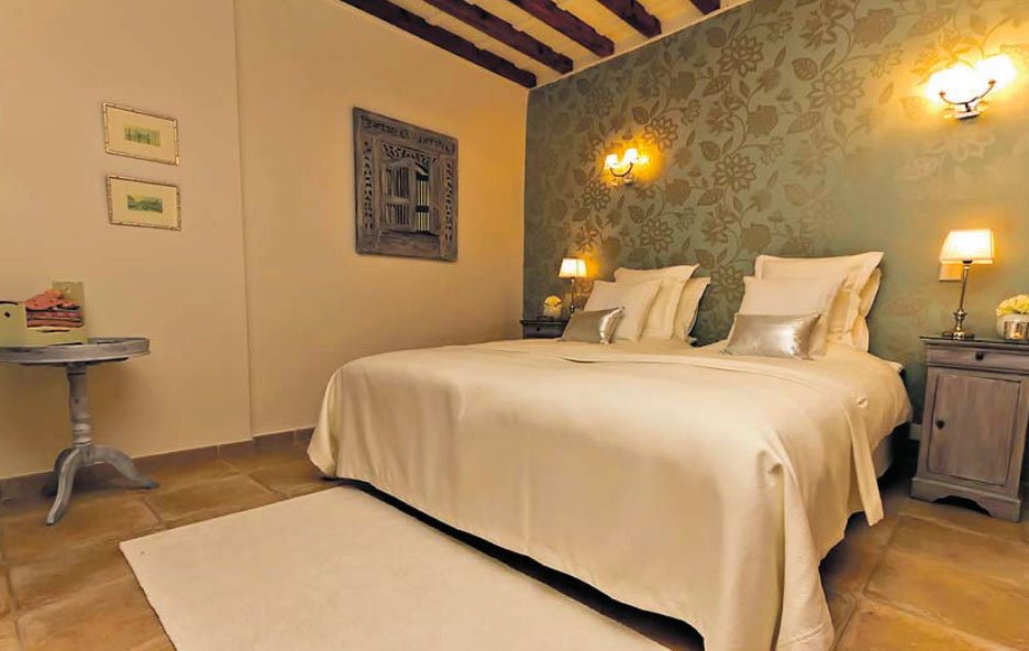 Dormitorio de San Feliu con cama doble, papel floral verde, vigas de madera y luz cálida