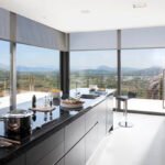 Cocina con isla de encimera negra, armarios oscuros y ventanales con estores y vistas a la montaña