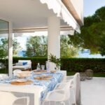 Terraza cubierta con toldo, mesa con mantel azul estampado y sillas blancas, seto y mar al fondo