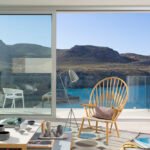 Butaca de madera y lámpara de pie ante un ventanal corredero con vistas a la cala