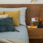 Detalle de la cama con cabecero de madera, cojines mostaza y verde azulado, mesita flotante y flores