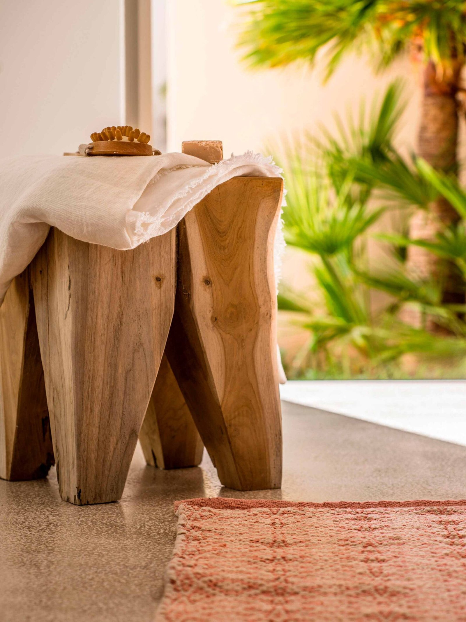 Detalle de taburete de madera natural con toalla blanca y alfombra rosa, con palmeras desenfocadas al fondo