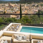 Terraza de estar con sofás de mimbre, piscina con tumbonas y panorama de tejados y montaña.
