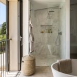 Ducha acristalada de mármol con rociador cromado, cesto de fibra y bañera blanca en primer plano.