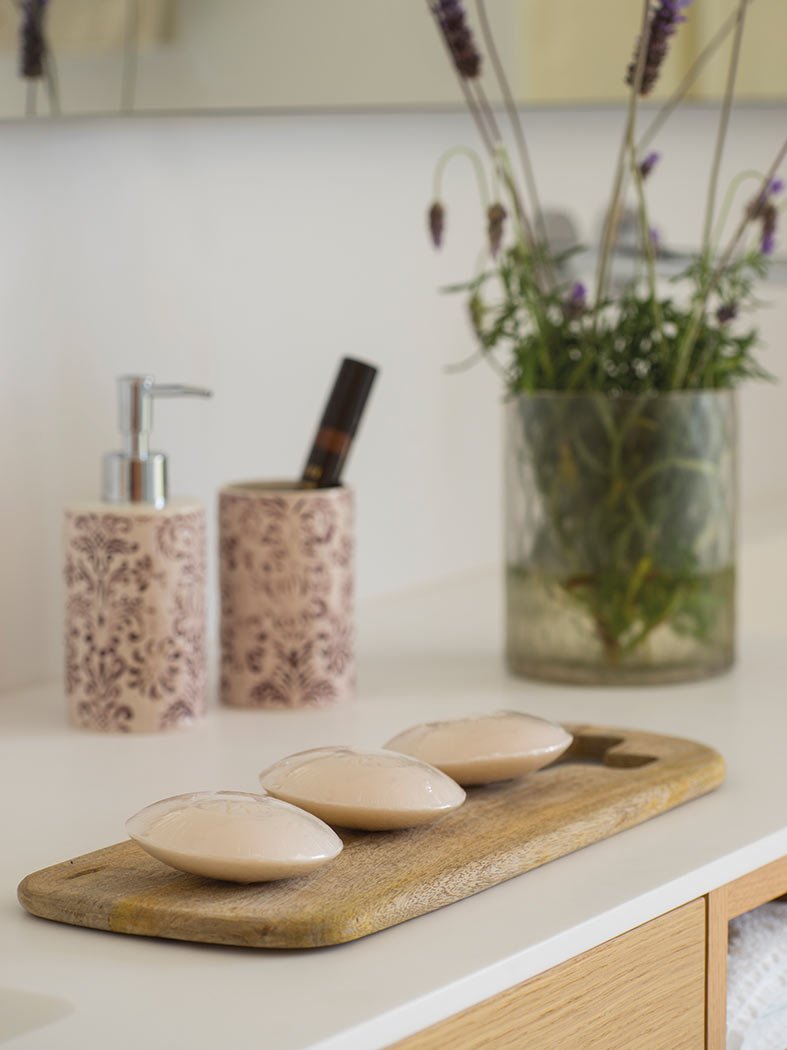 Detalle de jabones sobre tabla de madera, dosificador cerámico estampado y lavanda en un jarrón de cristal.
