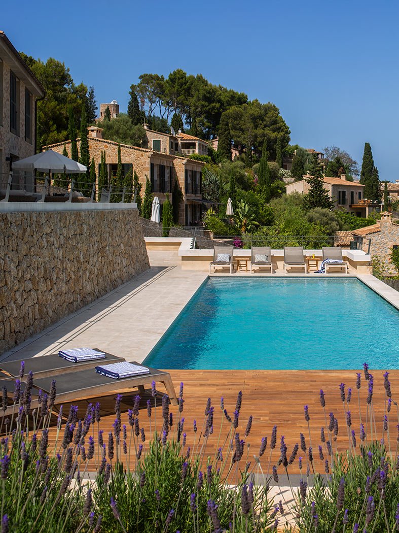 Piscina con tarima de madera, lavanda en primer plano, tumbonas, muro de piedra y casas al fondo.