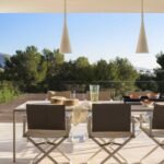 Comedor exterior con sillas plegables, mesa blanca, lámparas colgantes y pinos bajo un cielo azul
