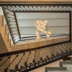 Escalera vista desde abajo con barandilla de forja, piedra clara y lámpara de esferas de cristal