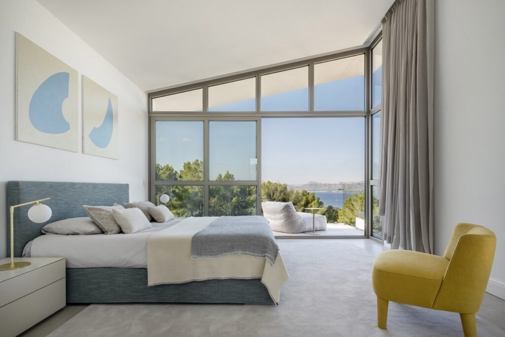 Dormitorio con cabecero azul grisáceo, sillón amarillo y gran ventanal con vistas al mar