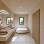 Baño con bañera exenta blanca, lavabo suspendido, espejo iluminado y ventana con vistas a árboles verdes