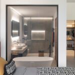 Dormitorio con cabecero rojo y baño en suite visible tras un cristal, con bañera exenta y doble lavabo