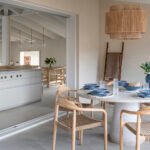 Comedor de terraza cubierta con mesa redonda blanca, sillas de madera y lámpara de fibra abierto a la cocina