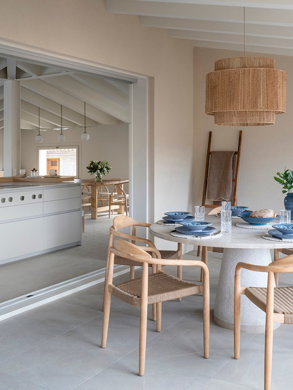 Comedor de terraza cubierta con mesa redonda blanca, sillas de madera y lámpara de fibra abierto a la cocina