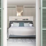 Dormitorio visto entre contraventanas verdes, con cama gris, cojines, cabecero con estante y suelo hidráulico