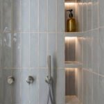 Detalle de ducha con azulejos verticales azul grisáceo, hornacinas iluminadas y grifería de acero